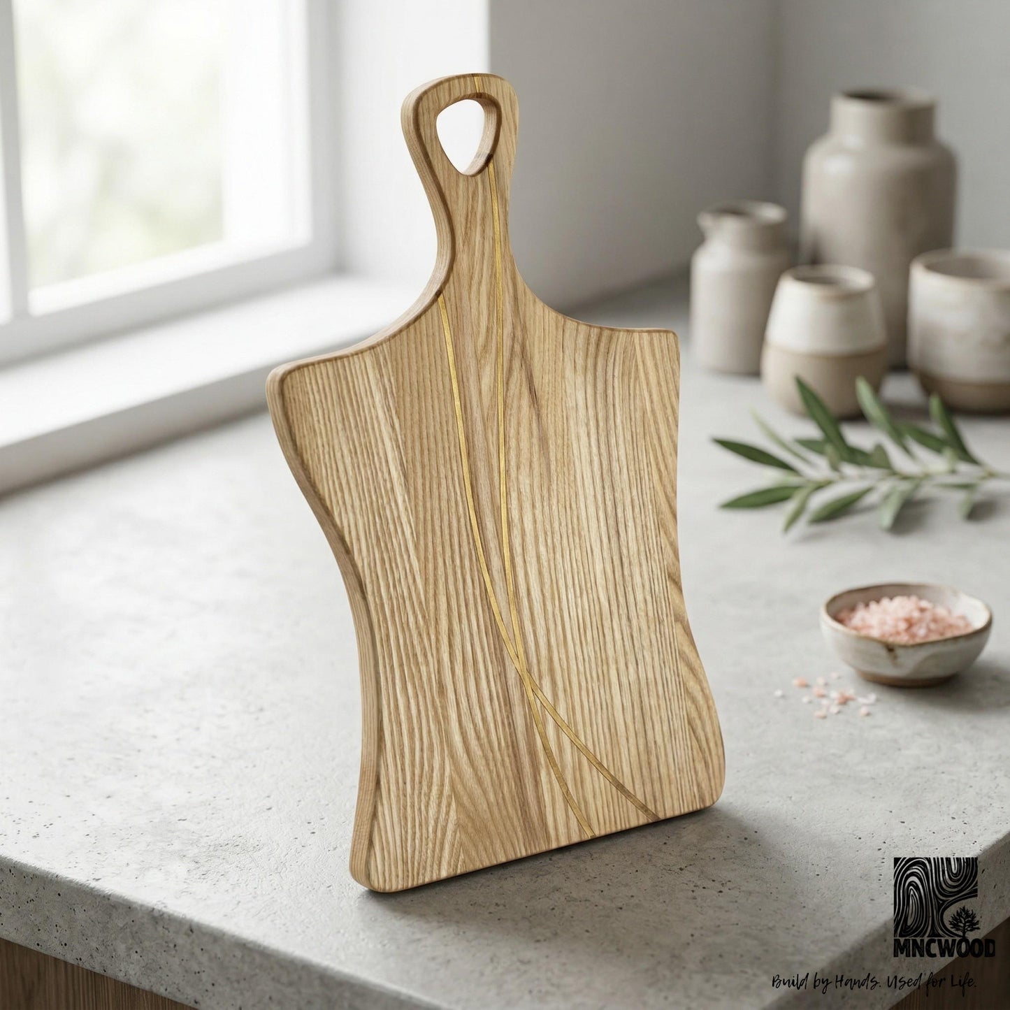 Wooden cutting board with handle on a kitchen counter with decorative vases and plants.